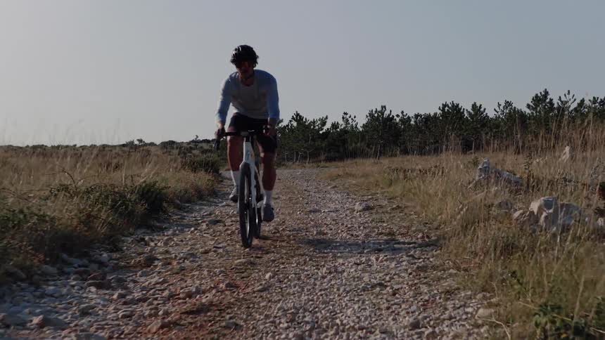 A male cyclist rides towards the camera on a rocky, gravel path through a dry, grassy landscape on a sunny day