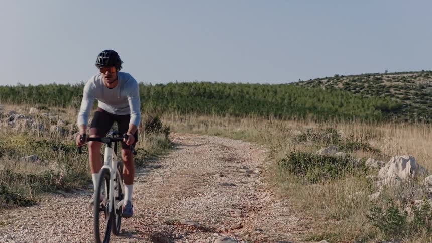 A male cyclist in athletic gear rides his gravel bike up a dirt path in the hilly countryside in slow motion