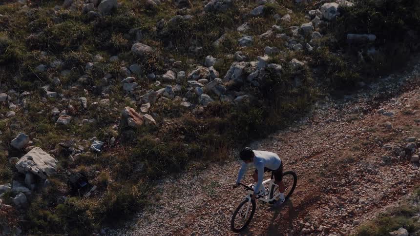 Top-down aerial shot follows a lone cyclist riding a mountain bike on a rugged gravel trail through a rocky landscape
