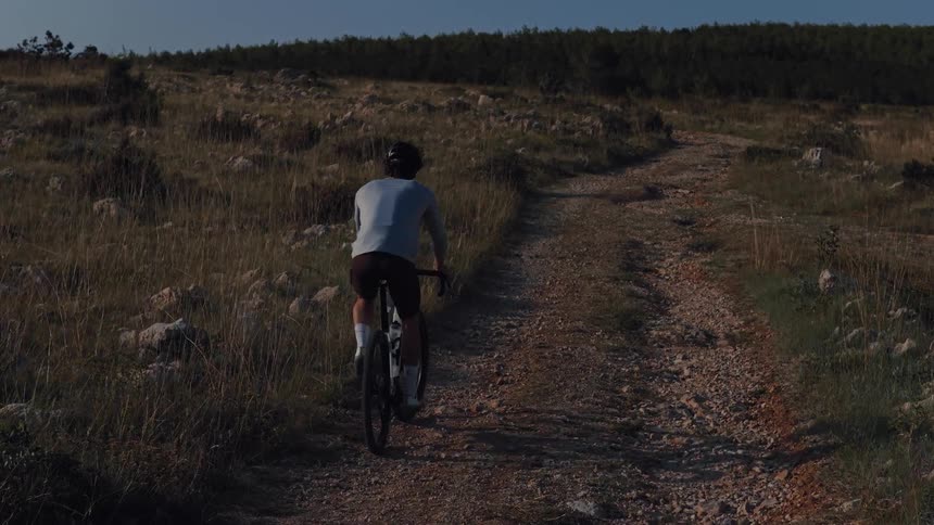 A male cyclist pedals in slow motion up a steep gravel road through a grassy, sunlit hill in the countryside