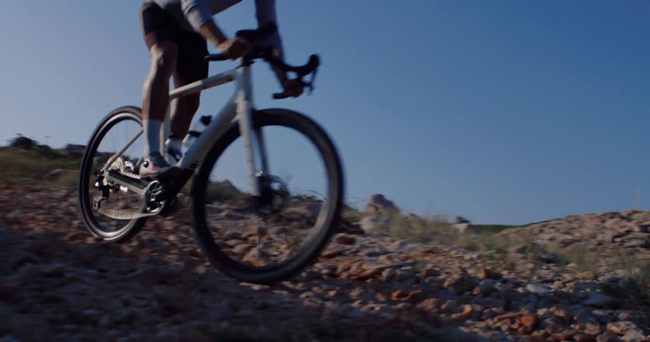 A slow-motion tracking shot follows a cyclist as they pedal uphill on a rugged, rocky trail during the golden hour