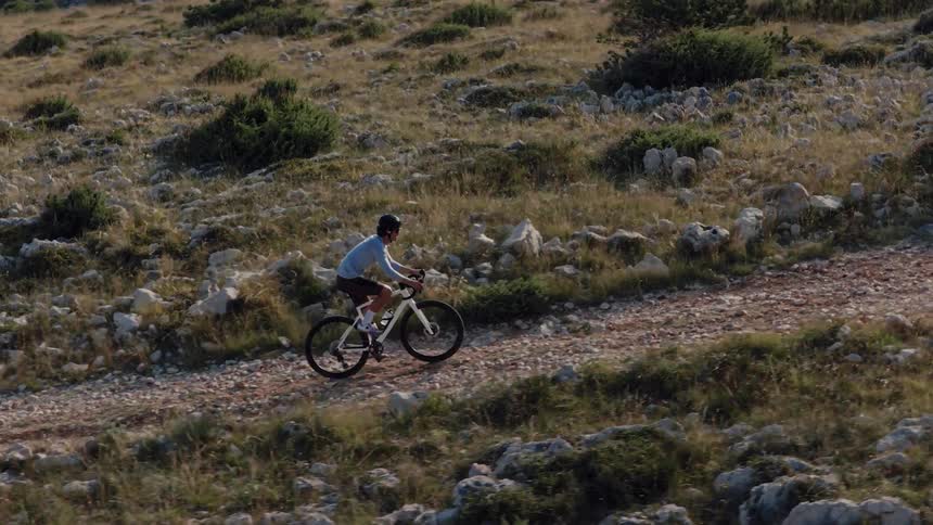 Slow motion side view of a cyclist pedaling up a steep gravel path on a rocky, sunlit mountain