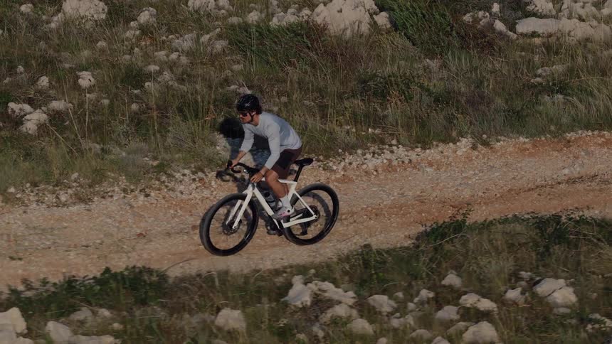 Slow-motion aerial shot of a cyclist pedaling up a rugged gravel path in a rocky, arid landscape