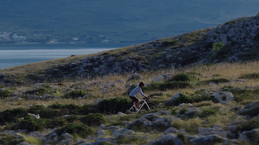 Aerial tracking shot of a cyclist navigating a rocky, sunlit trail on a hillside overlooking the sea