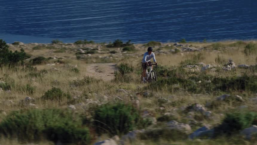 An aerial view shows a cyclist riding a gravel bike on a scenic coastal trail overlooking the blue sea