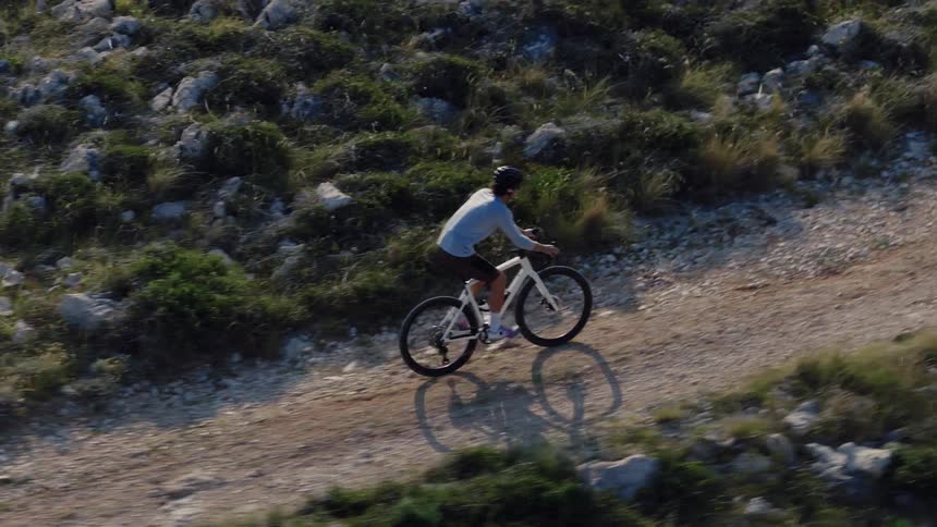 An aerial view tracks a man cycling uphill on a challenging, rocky mountain trail in slow motion