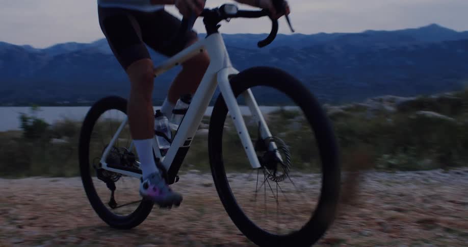 Slow motion tracking shot of a cyclist riding on a scenic gravel trail along the coast at dusk with mountains in the background