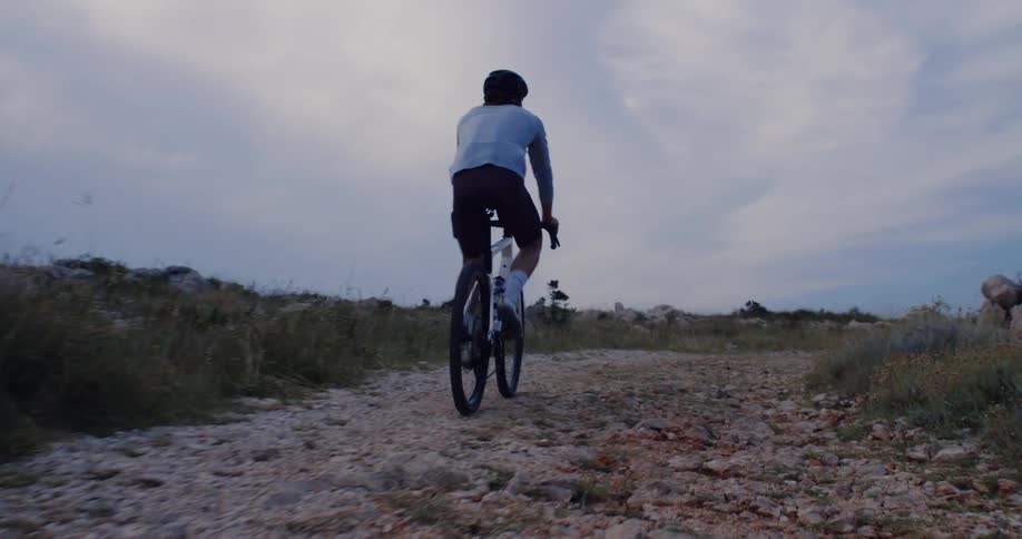Low-angle tracking shot of a cyclist riding uphill on a rugged gravel path during a moody, overcast evening