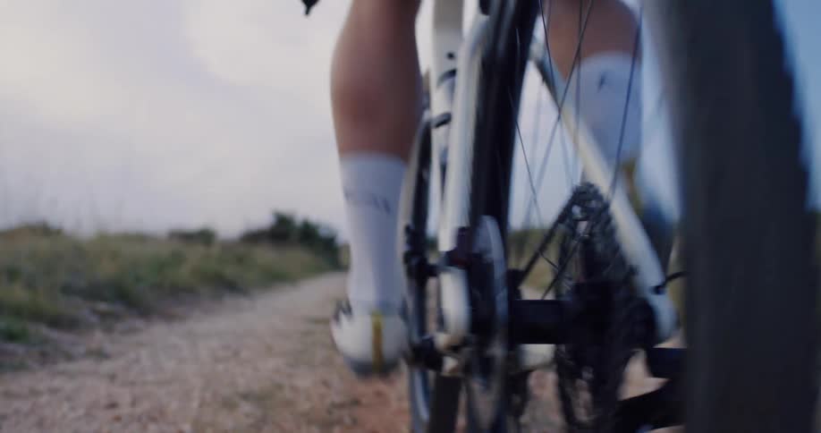 A detailed slow-motion close-up of a bicycle's rear wheel, cassette, and disc brake as a cyclist pedals along a gravel country road