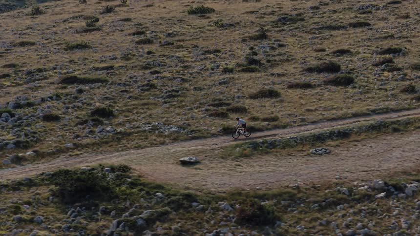High angle aerial shot of a lone mountain biker riding along a rugged dirt trail through a vast rocky landscape
