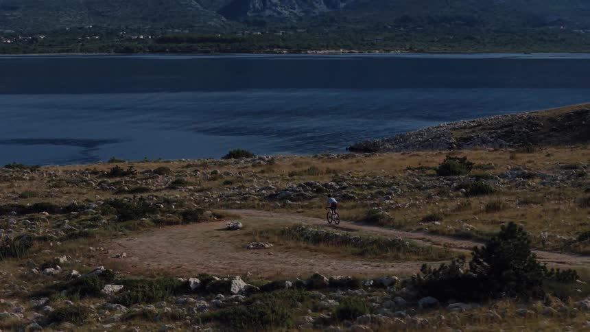 Aerial view of a mountain biker navigating a winding dirt trail on a rocky island coastline overlooking the ocean