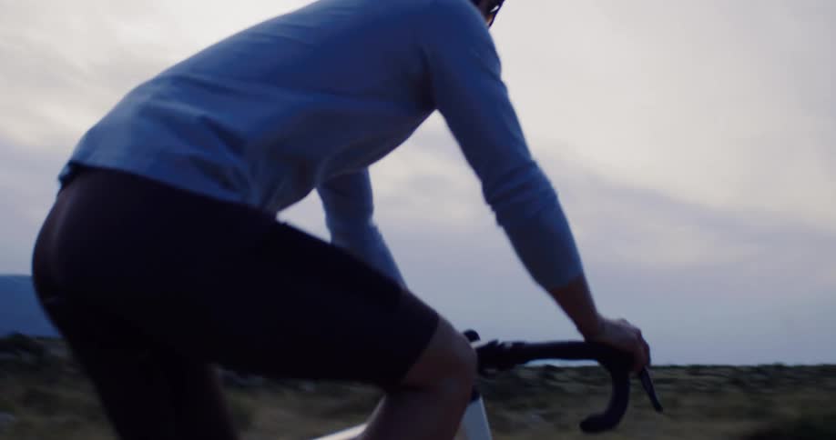A focused male cyclist in slow motion pedals his bike up a gravel mountain path with a scenic view of a lake and hills