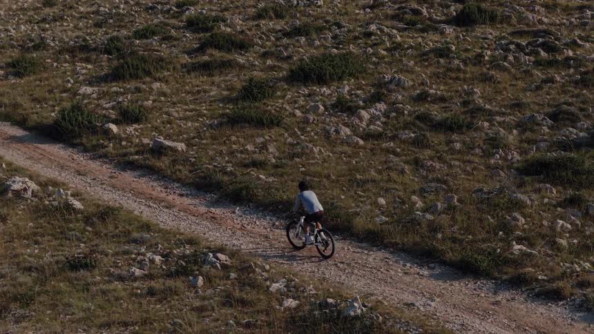 Slow-motion aerial shot follows a person mountain biking on a dirt road through a barren, rocky wilderness