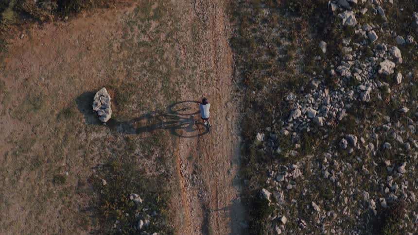 An aerial top-down view of a cyclist riding a gravel bike on a winding dirt path through a rocky, sunlit landscape