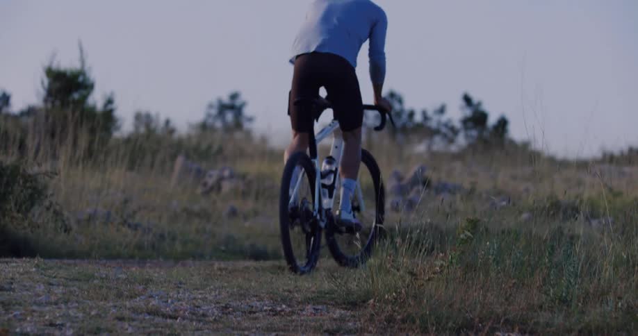 Slow motion shot of a cyclist riding away on a gravel path through a grassy field at dusk