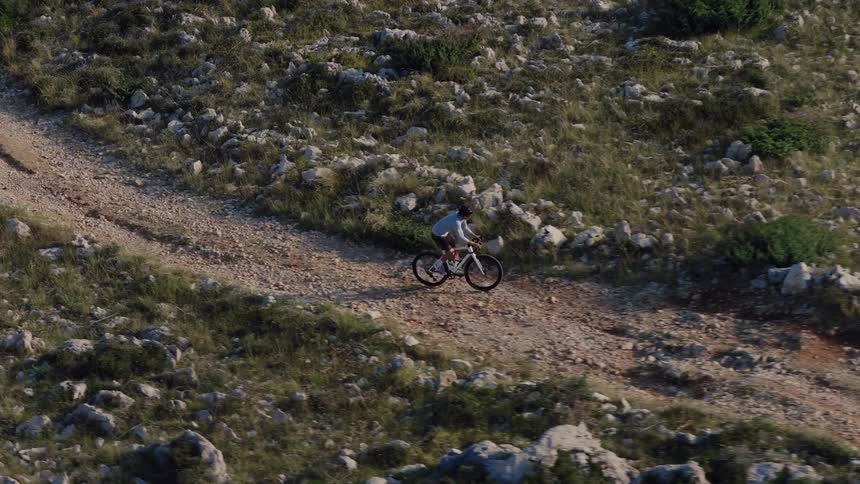 An aerial slow-motion shot captures a cyclist riding a mountain bike along a rugged dirt trail in a rocky, sun-drenched landscape