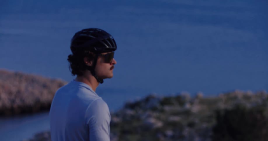 A male cyclist in a helmet and sunglasses takes a break to admire the serene ocean view from a rocky coastline as the shot comes into focus
