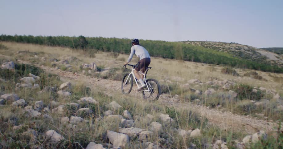 A male cyclist wearing a helmet rides his bike uphill on a rugged, rocky trail through a sun-drenched, grassy landscape in slow motion