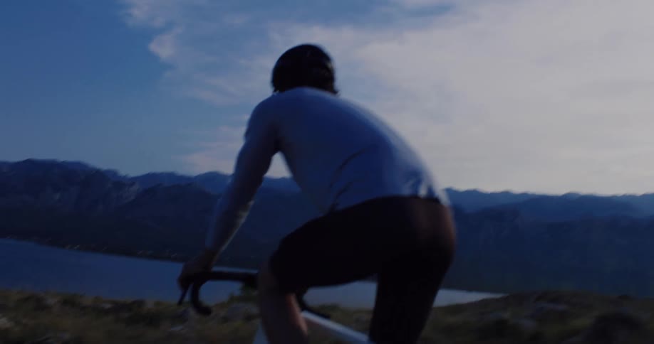 A determined cyclist rides his bike along a scenic mountain trail, with a stunning view of the water and distant mountains at dusk