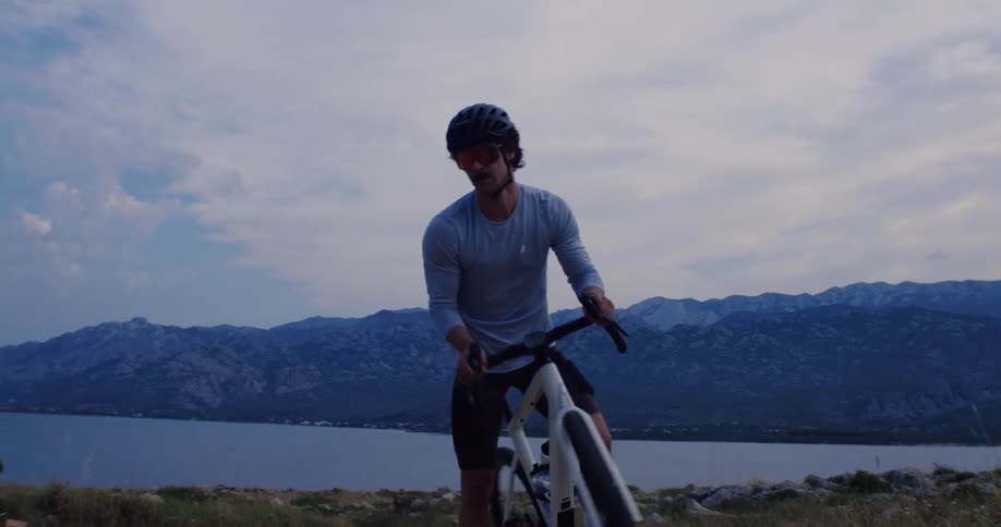 A male cyclist takes a moment to admire the dramatic mountain and sea landscape at dusk before continuing his ride on a gravel trail in slow motion