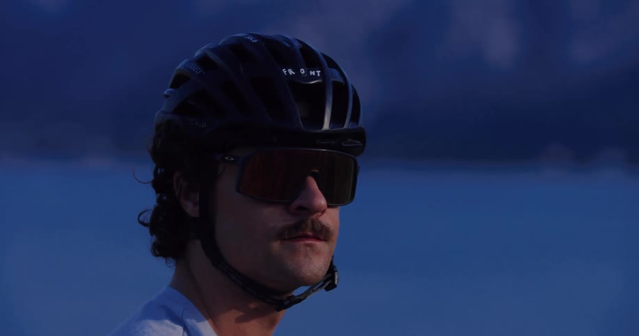 A slow-motion close-up profile of a male cyclist wearing a helmet and sunglasses, looking ahead with focus during an evening ride