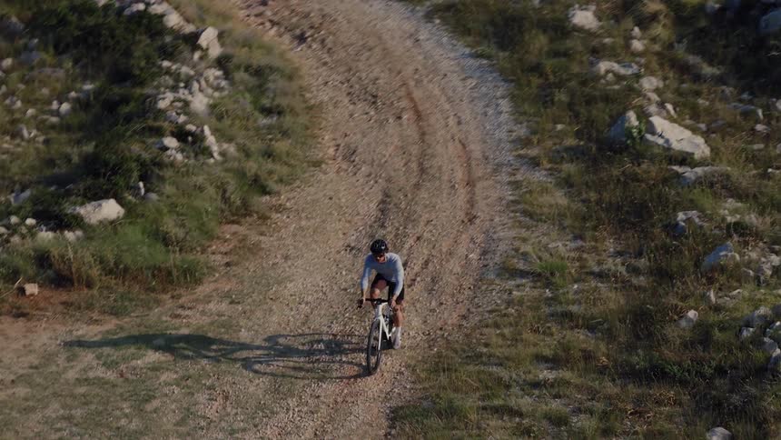 An aerial slow-motion shot follows a man riding his gravel bike along a rugged coastal path during the golden hour