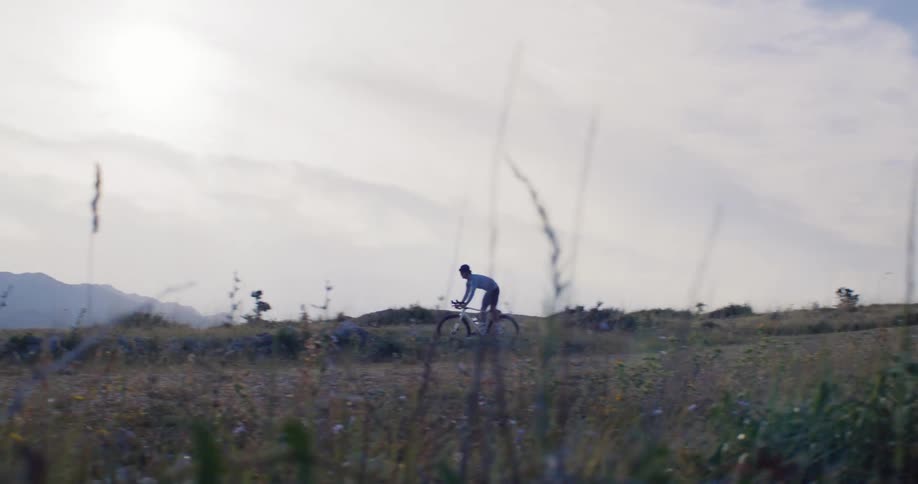 Slow motion footage shows a cyclist riding through a scenic, grassy landscape with mountains in the background