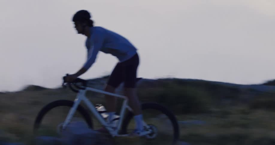 Slow motion tracking shot of a man cycling uphill on a rugged mountain trail at dusk