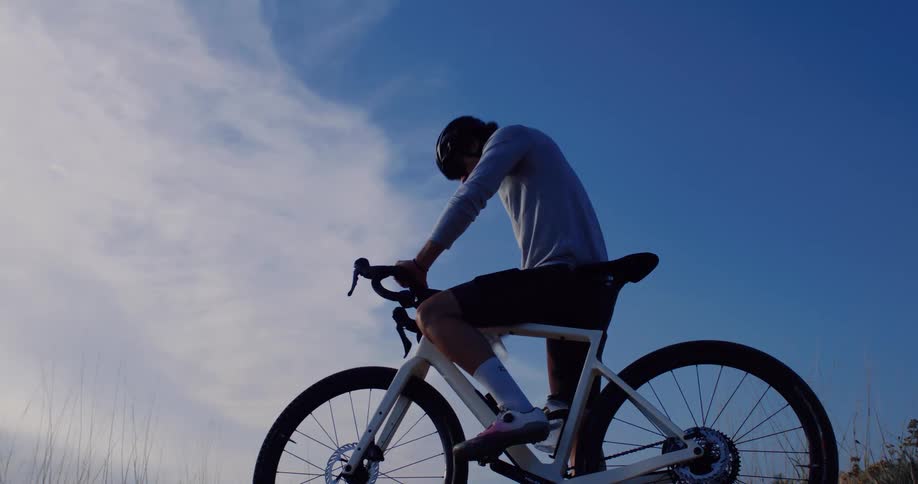In slow motion, a silhouetted cyclist takes a water break and then rides off against a clear blue sky