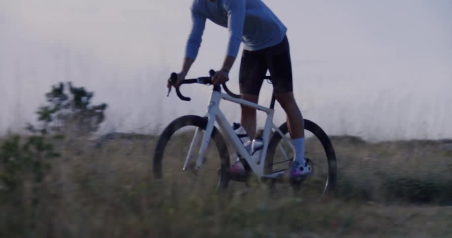 Slow motion side view of a cyclist riding his gravel bike uphill through a grassy field at dusk