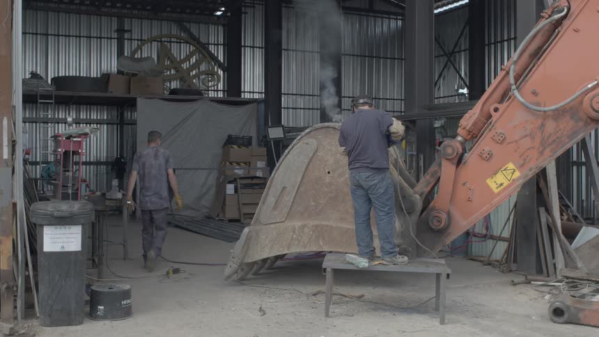 A welder repairs a large excavator bucket in an industrial workshop while another worker assists in the background