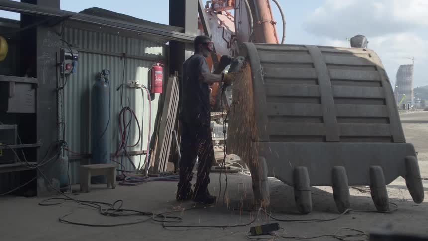 A metal worker uses an angle grinder to smooth the edge of a massive excavator bucket in a workshop, creating a shower of sparks