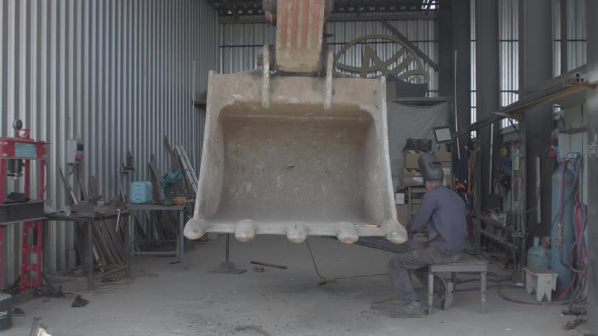 A professional welder repairs a large, suspended excavator bucket inside an industrial workshop