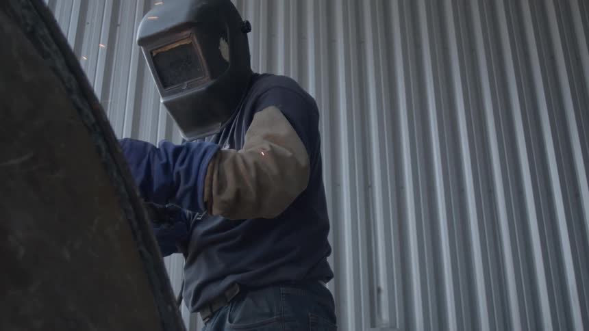 A welder in protective gear works on a piece of metal, with sparks flying in slow motion