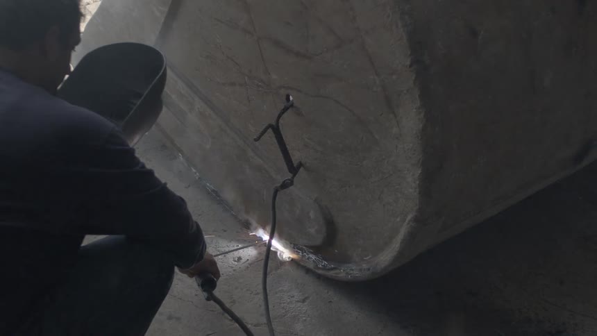 A welder wearing a protective mask repairs a large metal excavator bucket, creating bright sparks and smoke in a workshop