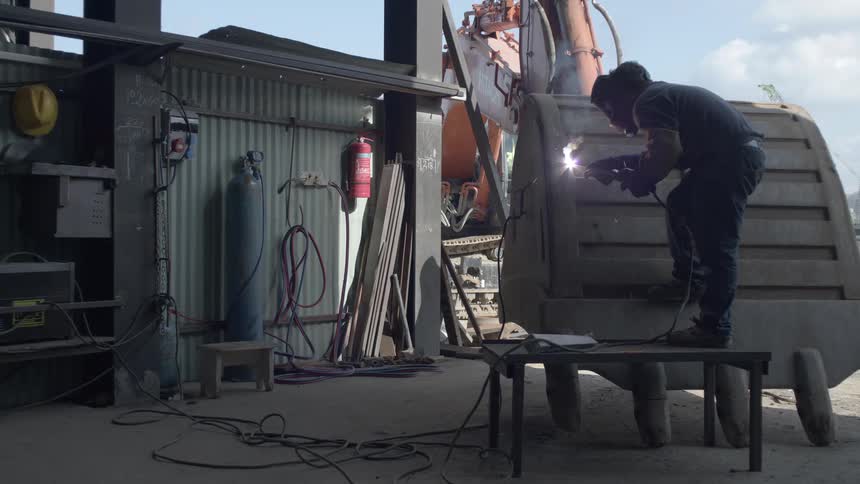 A skilled welder wearing a protective mask stands on a stool while repairing a massive excavator bucket in an industrial workshop
