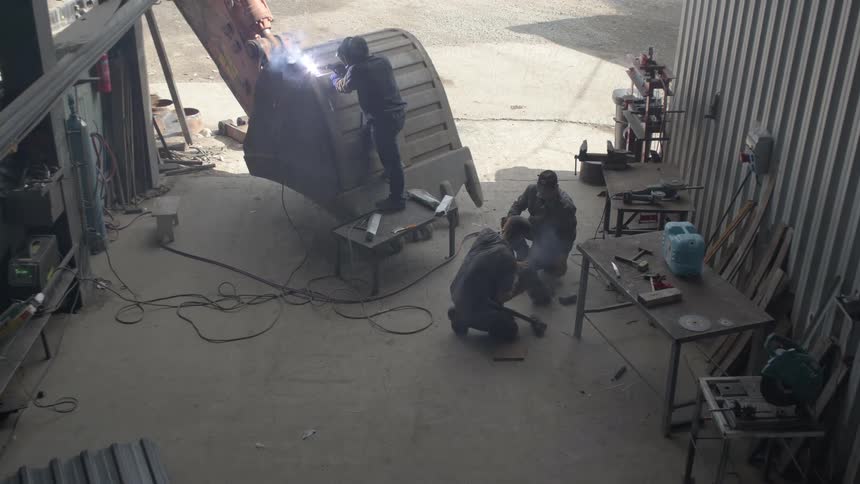 Three industrial workers weld and grind a massive excavator bucket during repairs in a metal fabrication shop