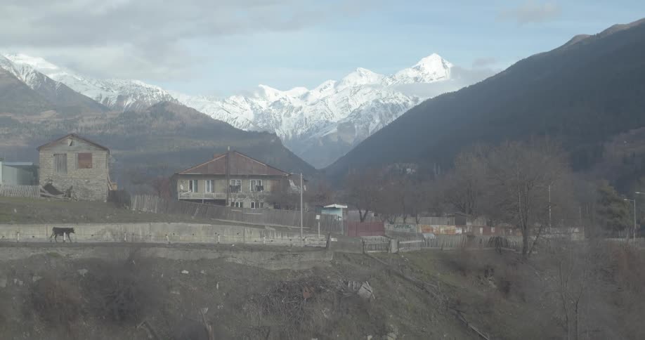 A quiet, rural village sits in a valley with majestic, snow-capped mountains towering in the distance