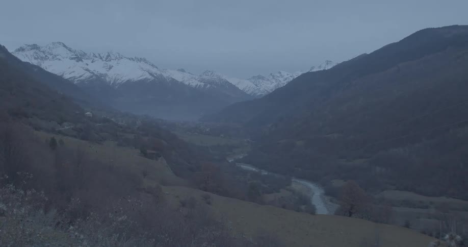 A majestic, moody landscape of a vast mountain valley with a river running through it and snow-capped peaks under an overcast sky
