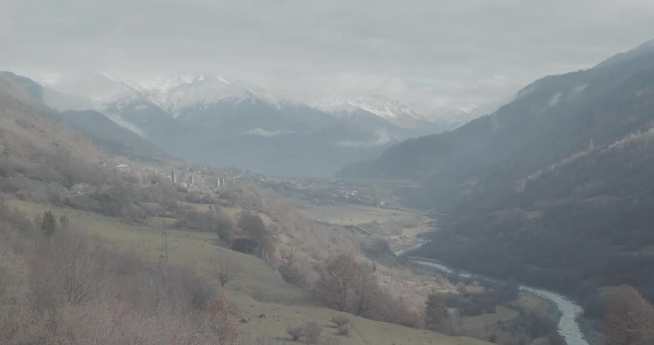 A wide, epic landscape of a misty mountain valley with a river winding through it and snow-capped peaks in the distance