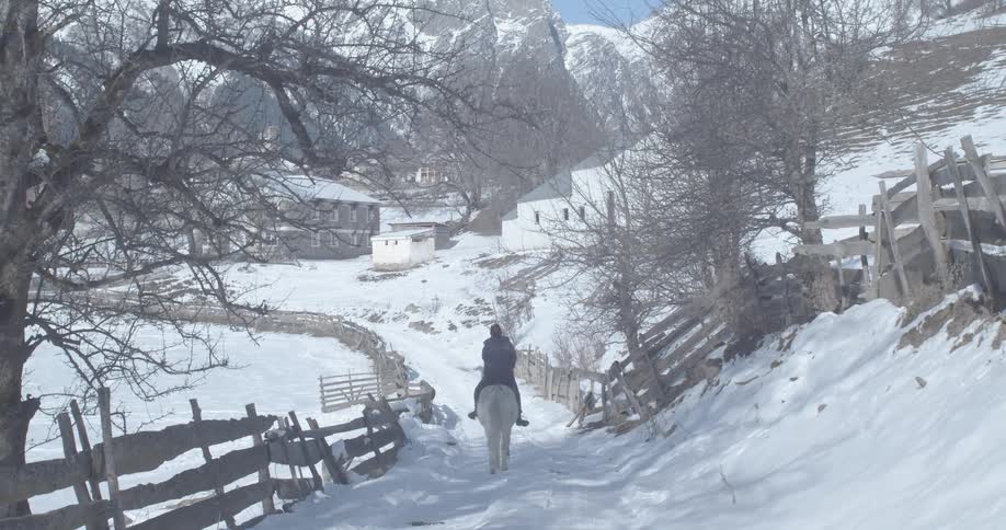 A lone equestrian on a white horse travels down a snow-covered country road towards a remote village nestled in the mountains