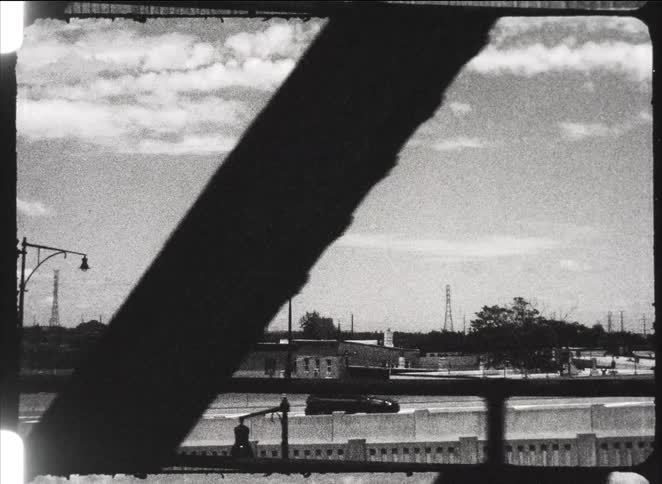 Gritty, black and white archival footage shows a highway and industrial landscape from the window of a moving vehicle