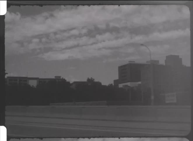 Vintage black and white footage shows a view of the city skyline and clouds from a moving car on the highway