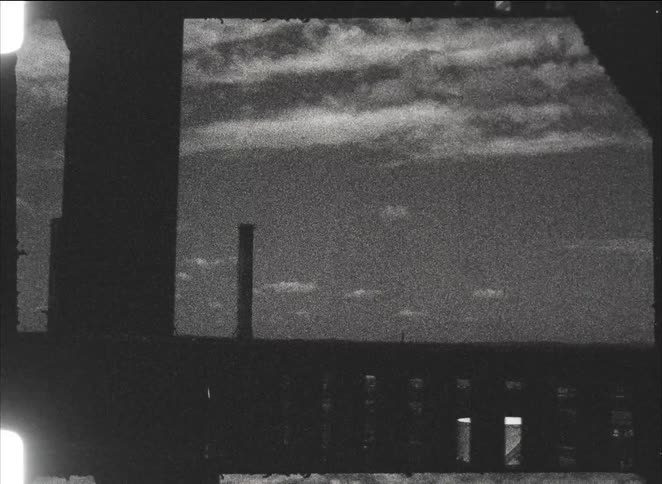 A grainy, black and white archival view of a silhouetted industrial skyline with smokestacks seen from a moving vehicle