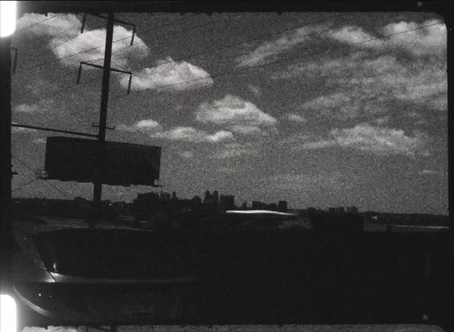 A grainy, black and white vintage film shot from a moving vehicle captures a distant city skyline under a cloudy sky