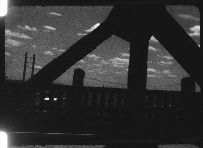 Vintage black and white 8mm footage captures a grainy view of a cloudy sky while driving across a steel truss bridge