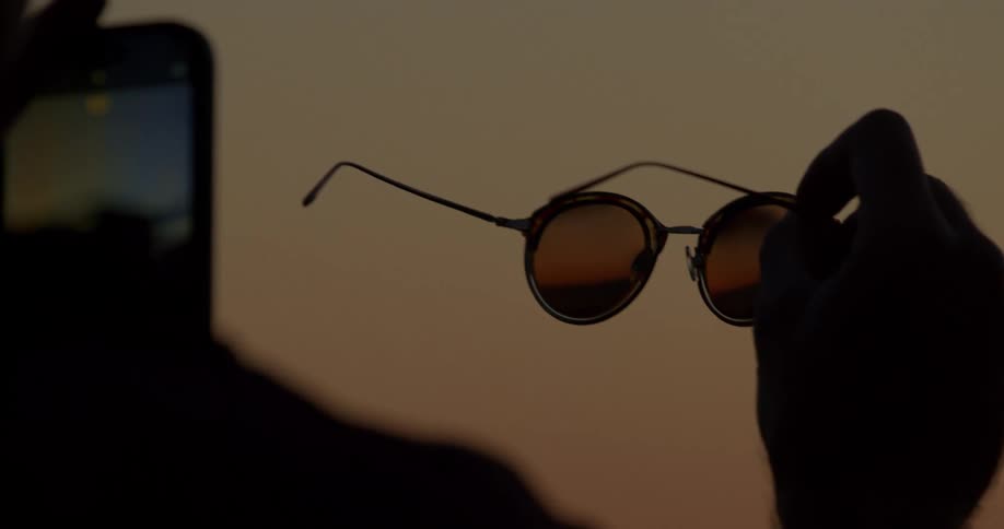 In slow motion, a silhouetted person holds up a pair of sunglasses reflecting the sunset, while another person captures the artistic moment on their phone