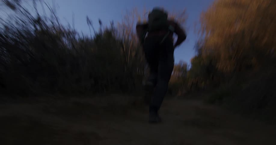 A low-angle action shot follows a person with a backpack running frantically up a dark, dusty trail at dusk