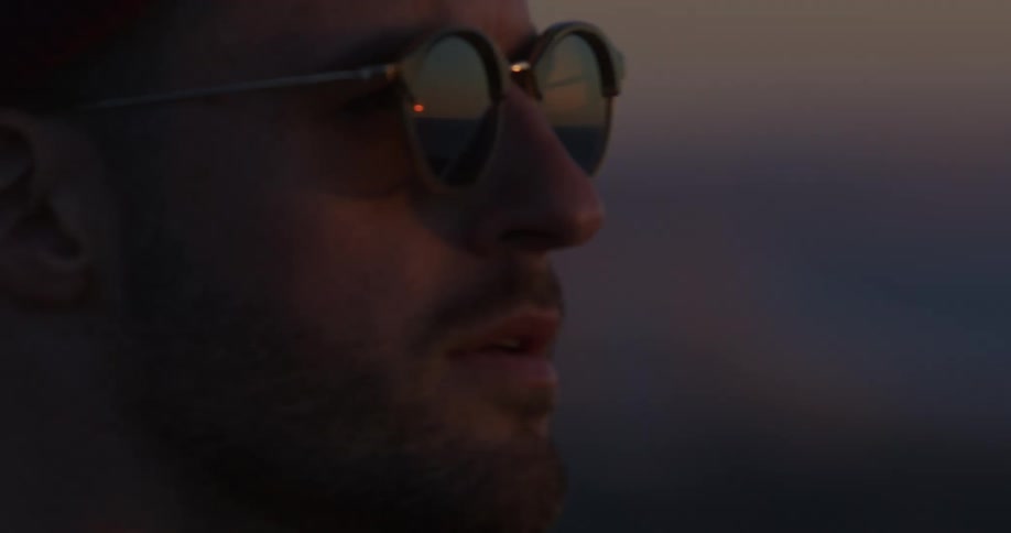 A man watches the sunset over a coastal city, with the beautiful view reflected in his sunglasses as the focus shifts to him