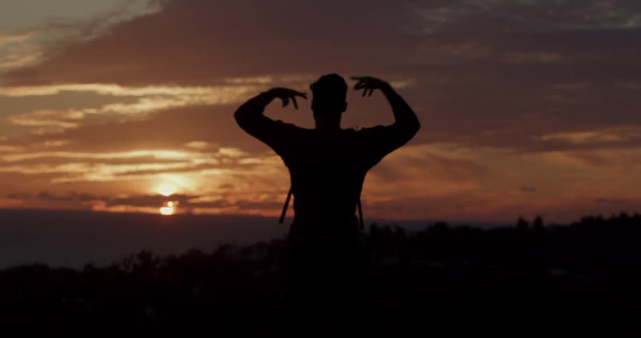 A silhouetted man performs a peaceful, meditative stretch while watching the sunset over the ocean